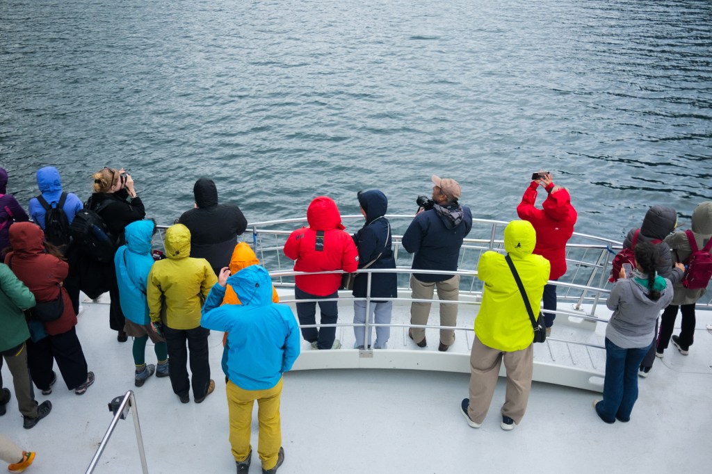 A group of people wearing colorful jackets observing water from the deck of a boat, some taking photos with cameras and phones.