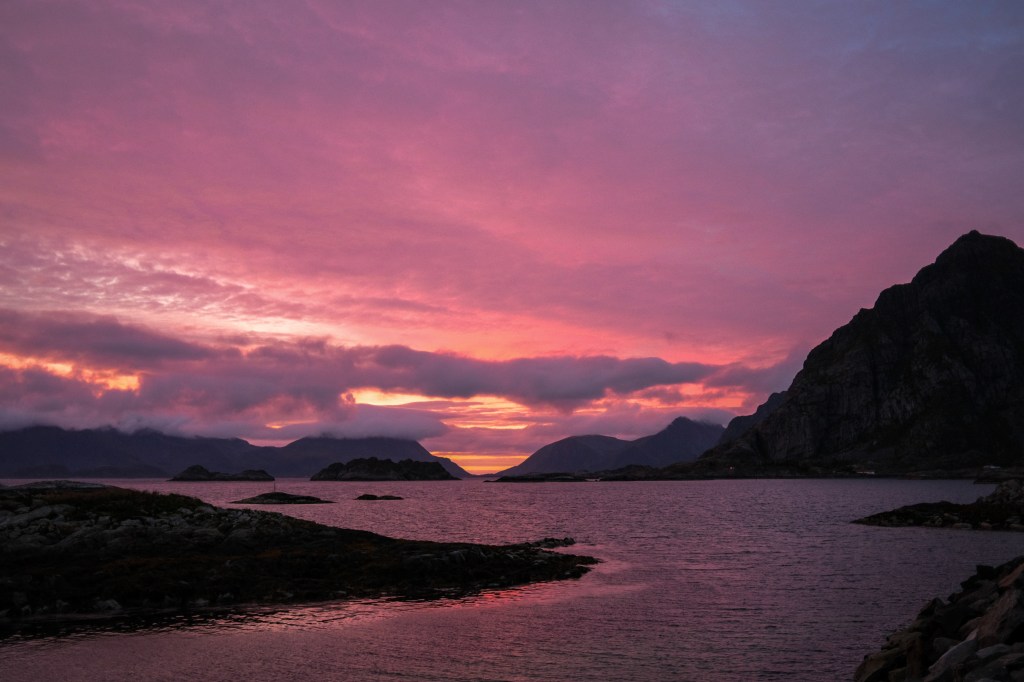 A stunning sunset over a tranquil sea, with dramatic purple and pink clouds illuminating the sky and silhouette of mountains in the background.