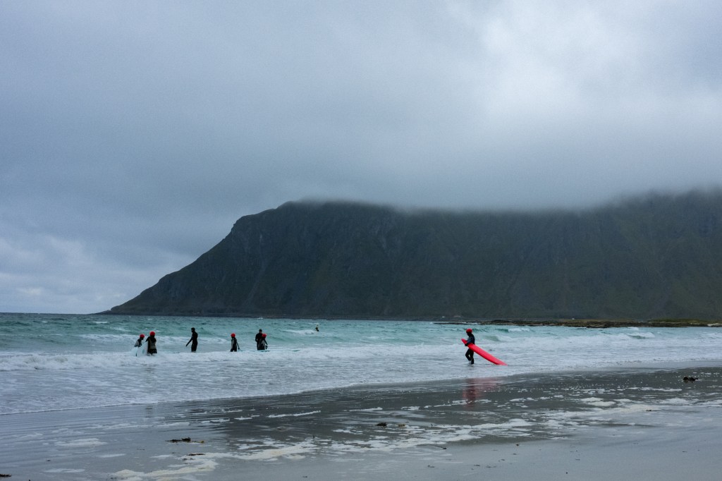 Surfers in wetsuits walking along a beach with waves, a mountain covered in clouds in the background.