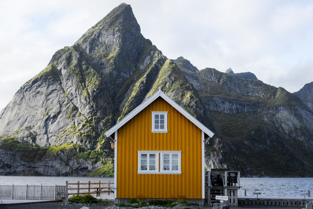 A bright yellow cabin situated by a body of water, with towering rocky mountains in the background under a cloudy sky.