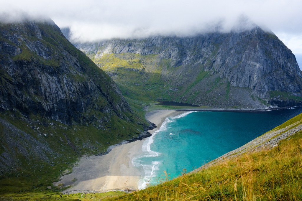 Aerial view of a secluded beach surrounded by rocky cliffs and green hills, with turquoise water and a cloudy sky above.