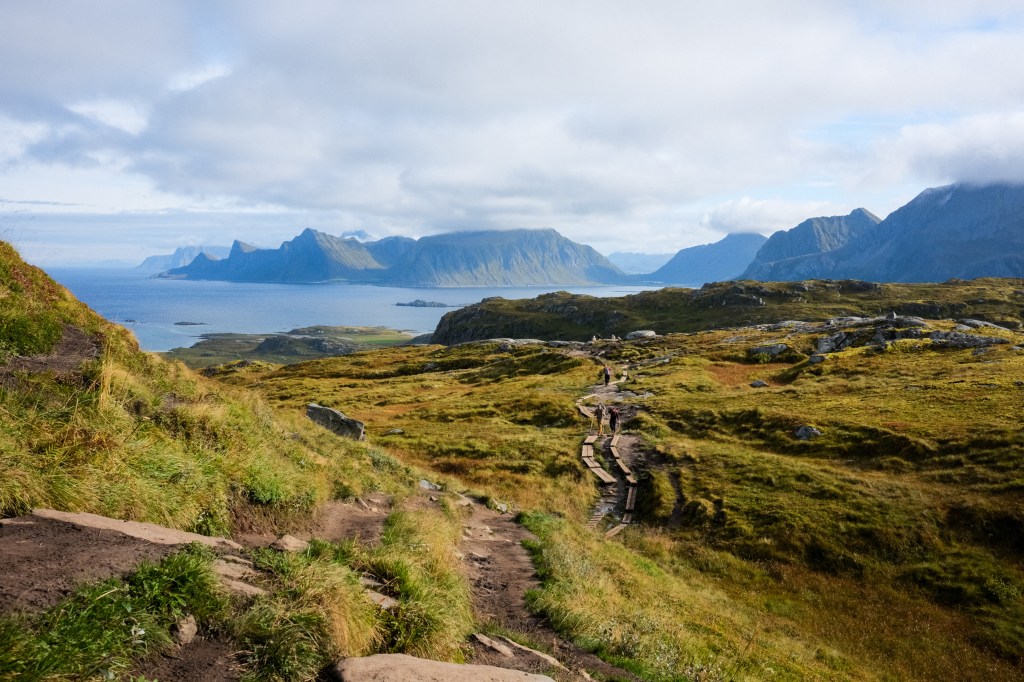 A scenic view of a hilly landscape with a pathway leading through lush green grass, overlooking a tranquil sea and distant mountains under a partly cloudy sky.