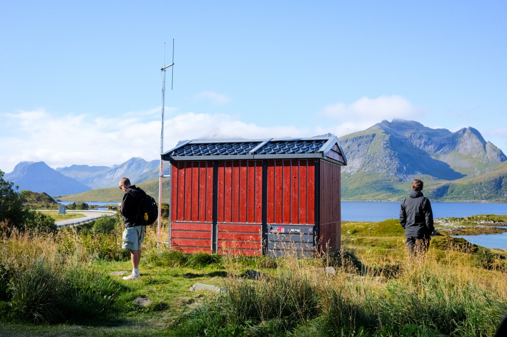 Two people standing near a small red cabin with a mountain backdrop and a body of water in the distance, under a clear blue sky.