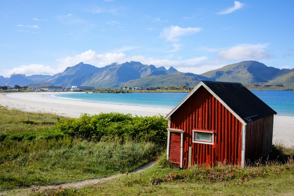 A picturesque beach scene featuring a small red wooden cabin near a sandy shore, with turquoise waters and distant mountains under a blue sky.