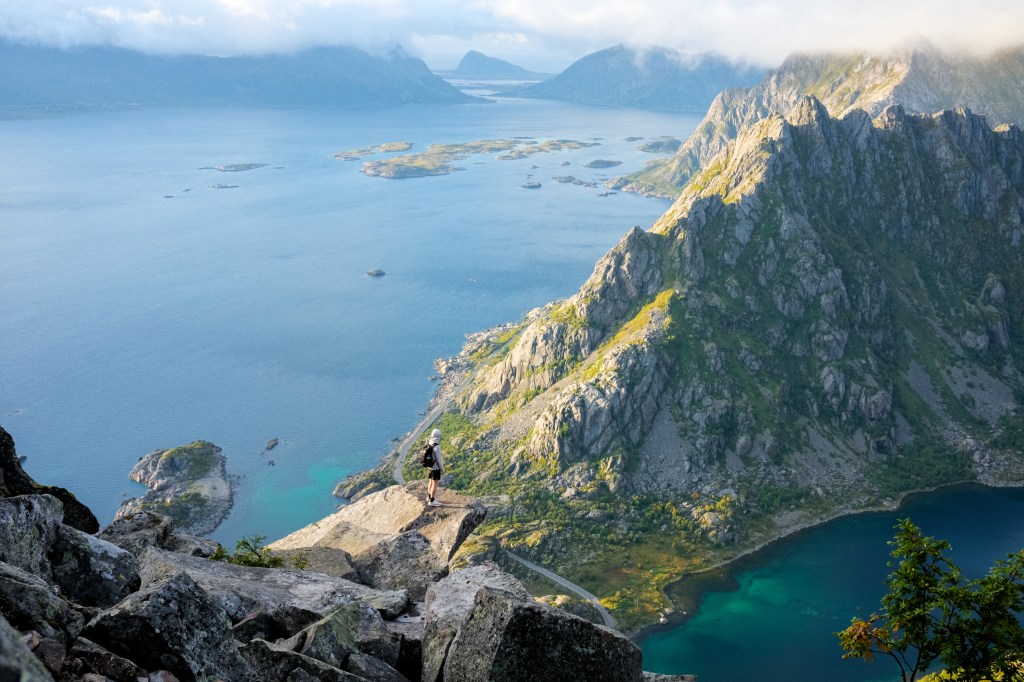A person standing on a rocky outcrop overlooking a stunning fjord with islands and mountains in the background, illuminated by sunlight under a partly cloudy sky.