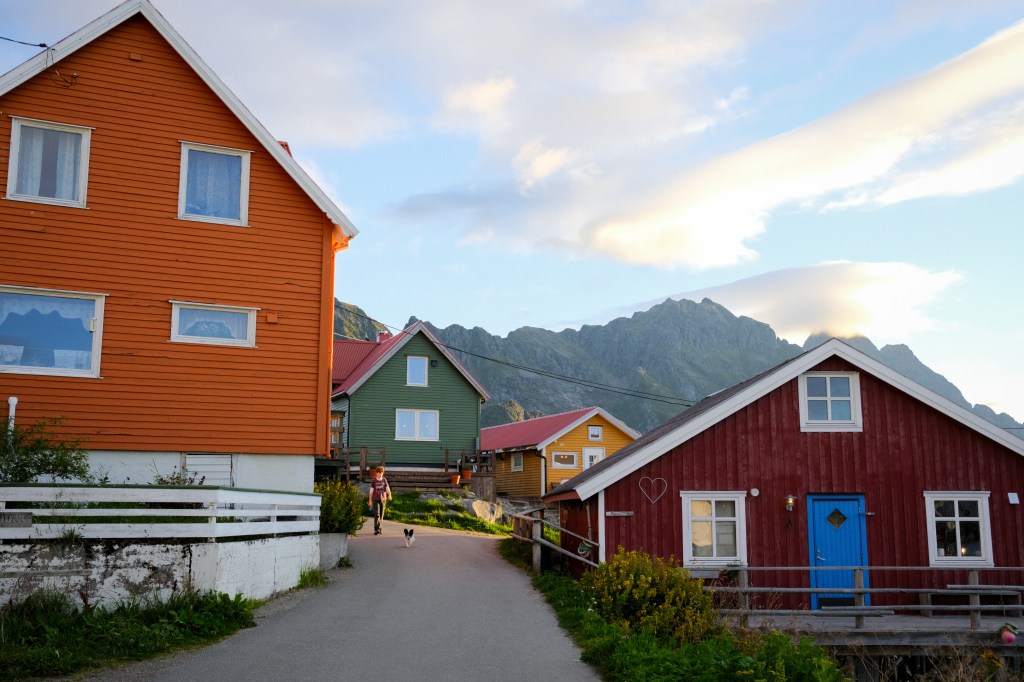 Colorful wooden houses along a pathway with a mountain backdrop and a person walking a dog.