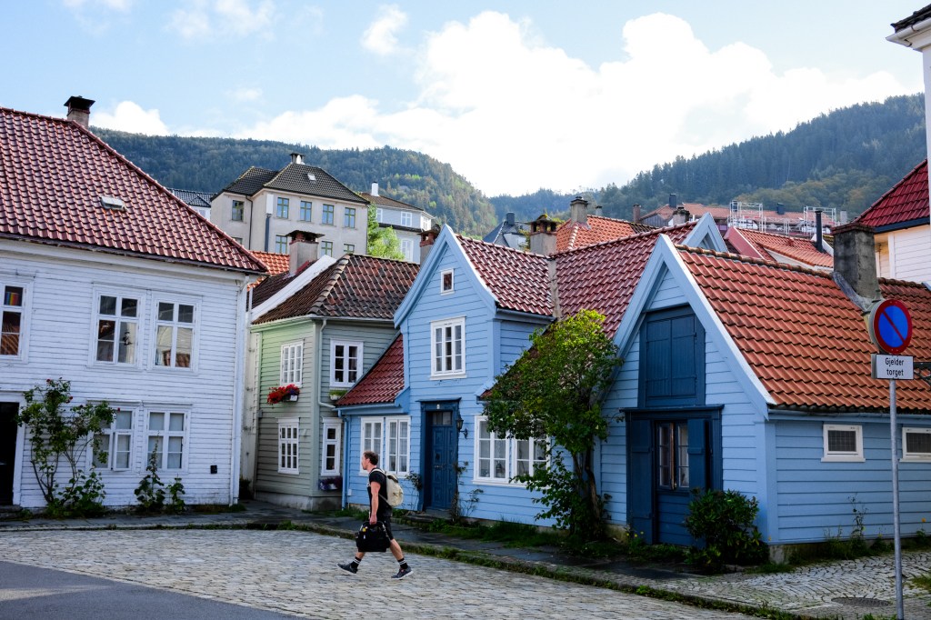 A street scene featuring colorful, historical houses with red rooftops, including white and blue buildings, set against a backdrop of green hills and a blue sky. A person walks down the cobblestone path near the houses.
