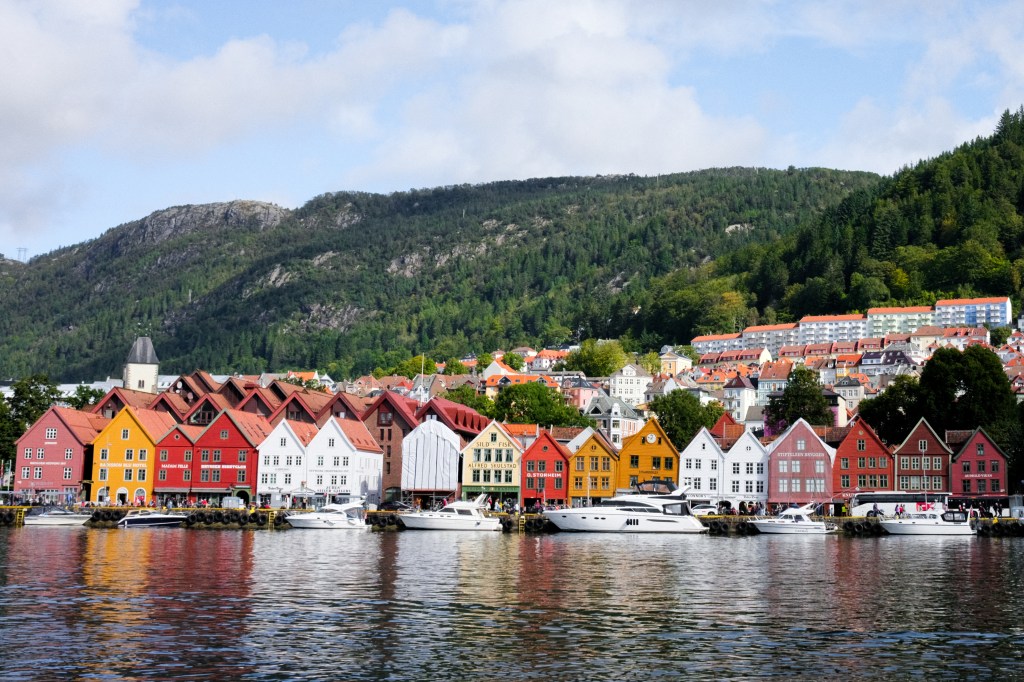 Colorful waterfront buildings along a harbor with boats, surrounded by lush green hills under a partly cloudy sky.