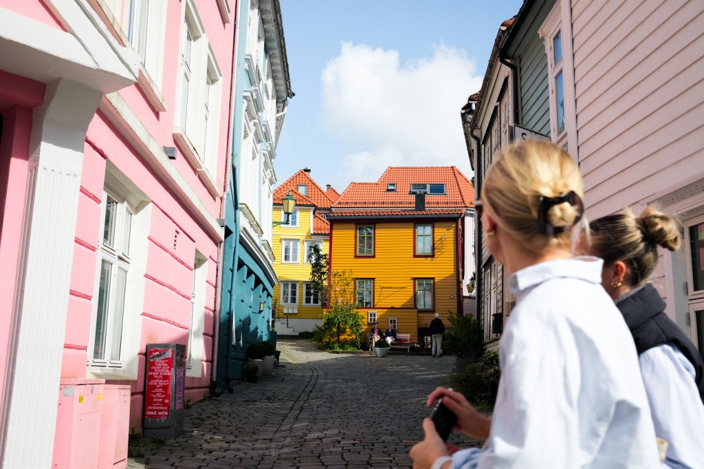 A scenic cobblestone street lined with colorful buildings, including pink, teal, and yellow houses, under a partly cloudy sky. Two people in the foreground are admiring the view.