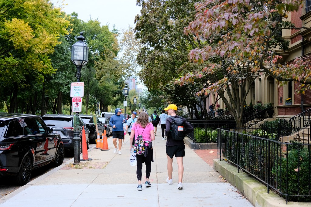 Two people walking on a tree-lined sidewalk surrounded by parked cars, with a few pedestrians visible in the background.