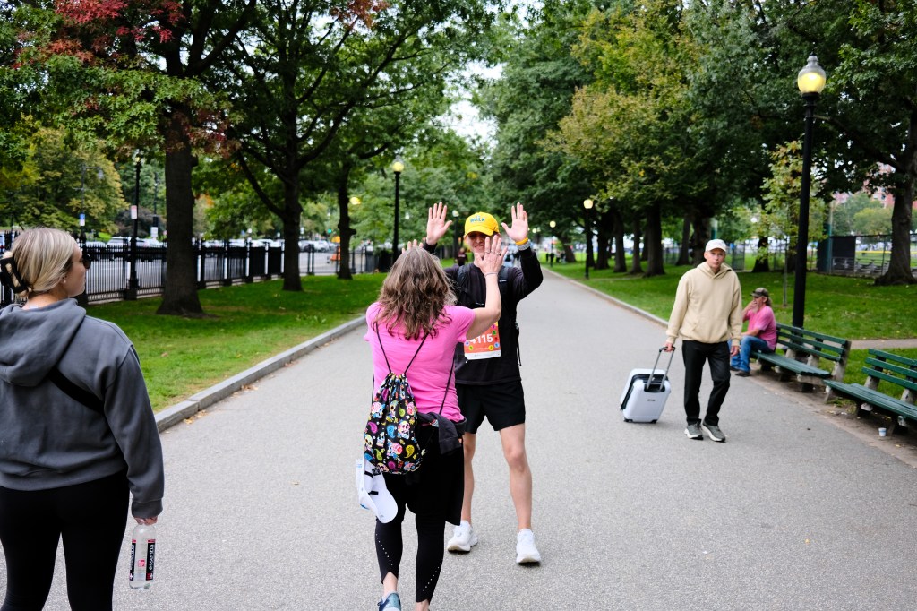 Two people interacting in a park setting; one is waving while the other approaches from behind. Several trees and park benches are visible.