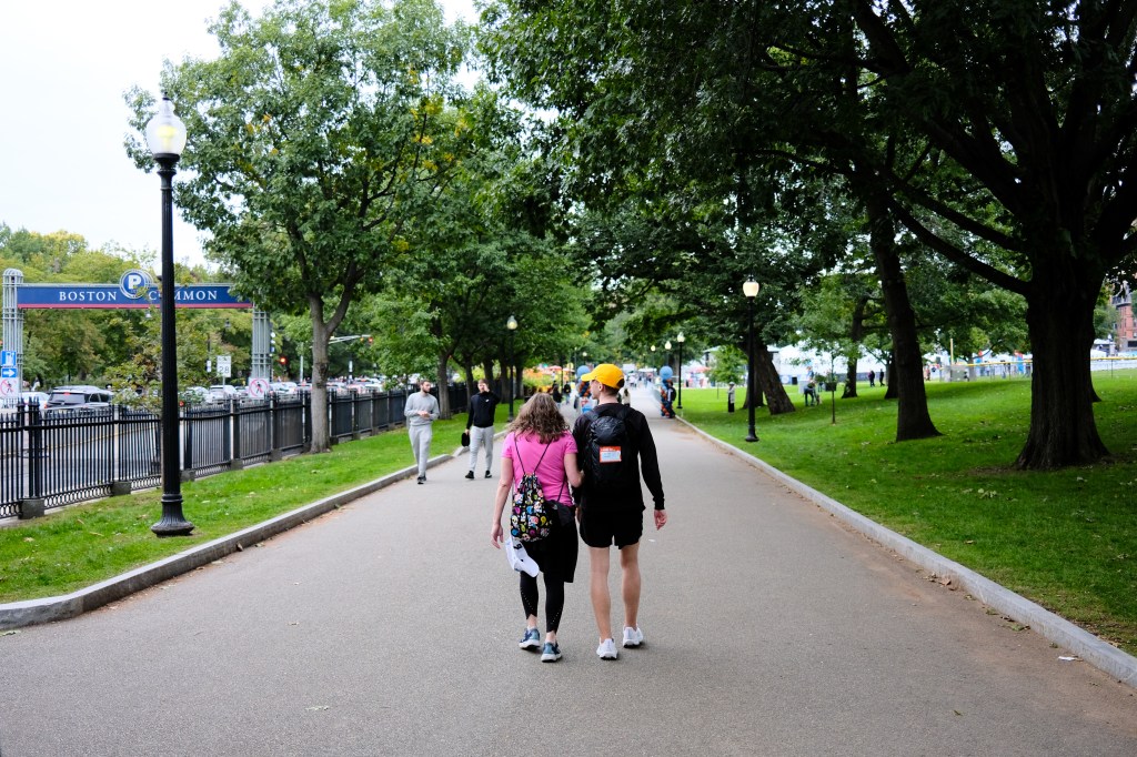 A couple walks hand in hand along a path in Boston Common, surrounded by trees and greenery, with people in the background.