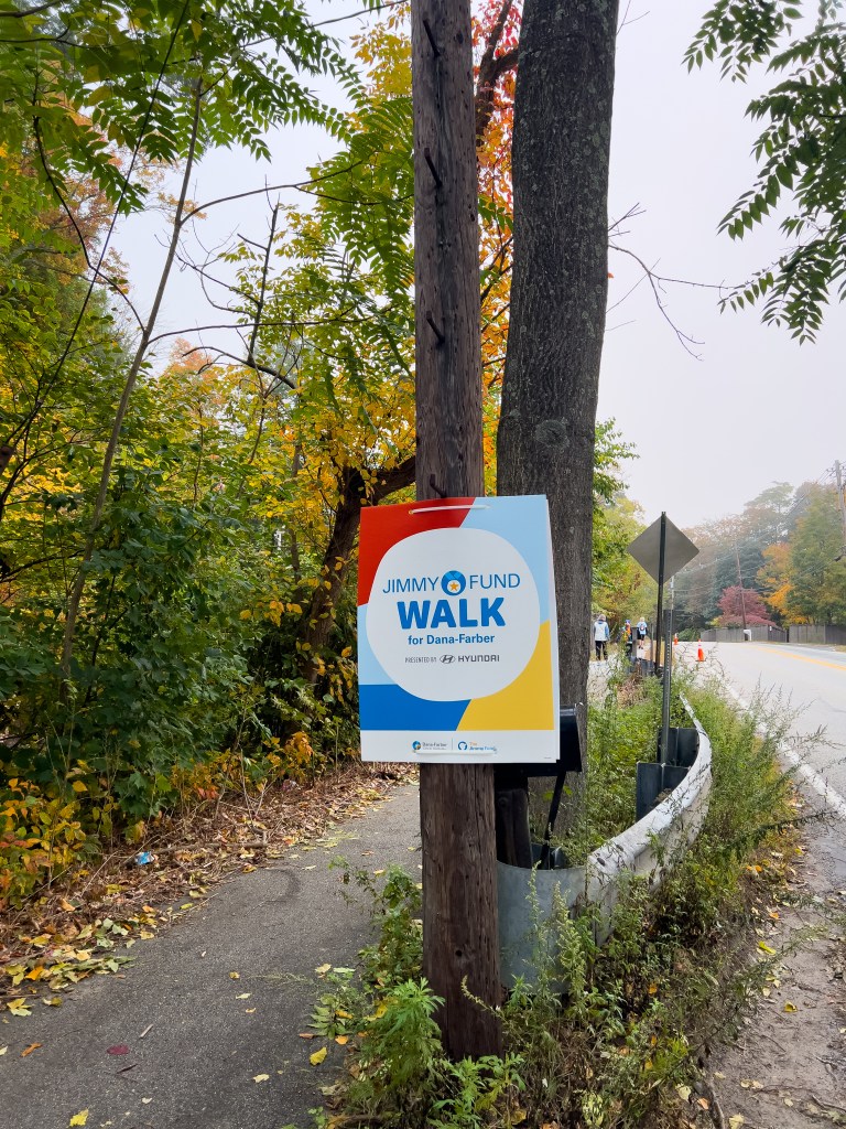 Sign for the Jimmy Fund Walk for Dana-Farber beside a path surrounded by autumn foliage.