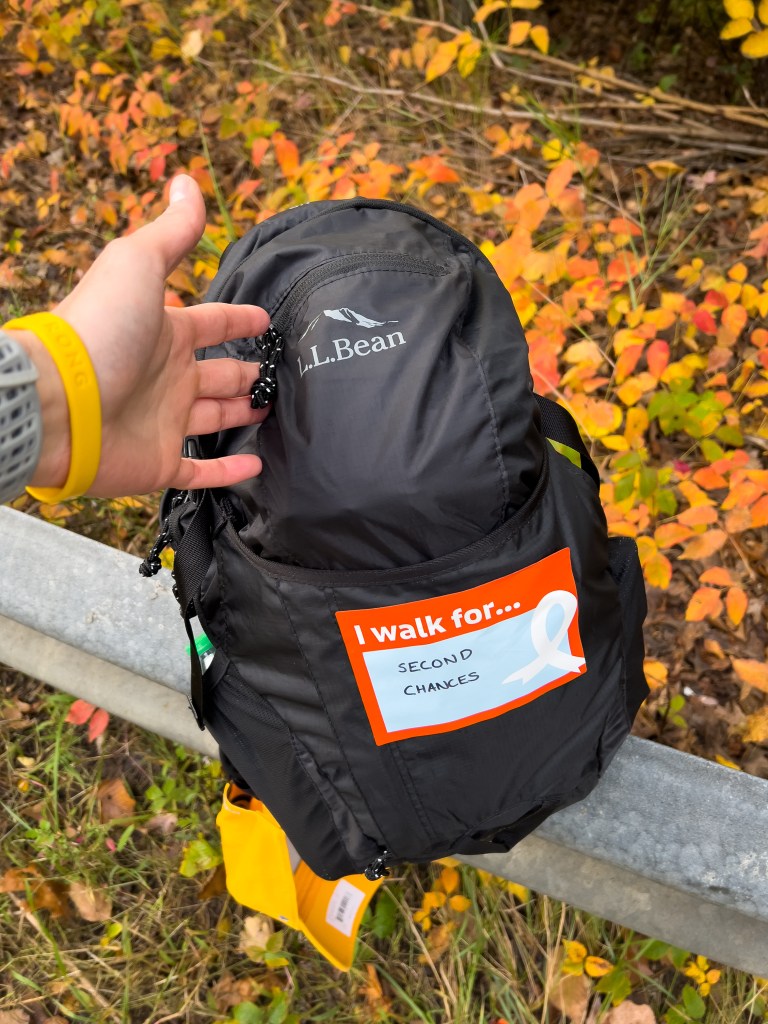 A hand holding a black L.L. Bean backpack with an orange sticker that reads 'I walk for... SECOND CHANCES.' The background features autumn leaves in various shades of orange and yellow.