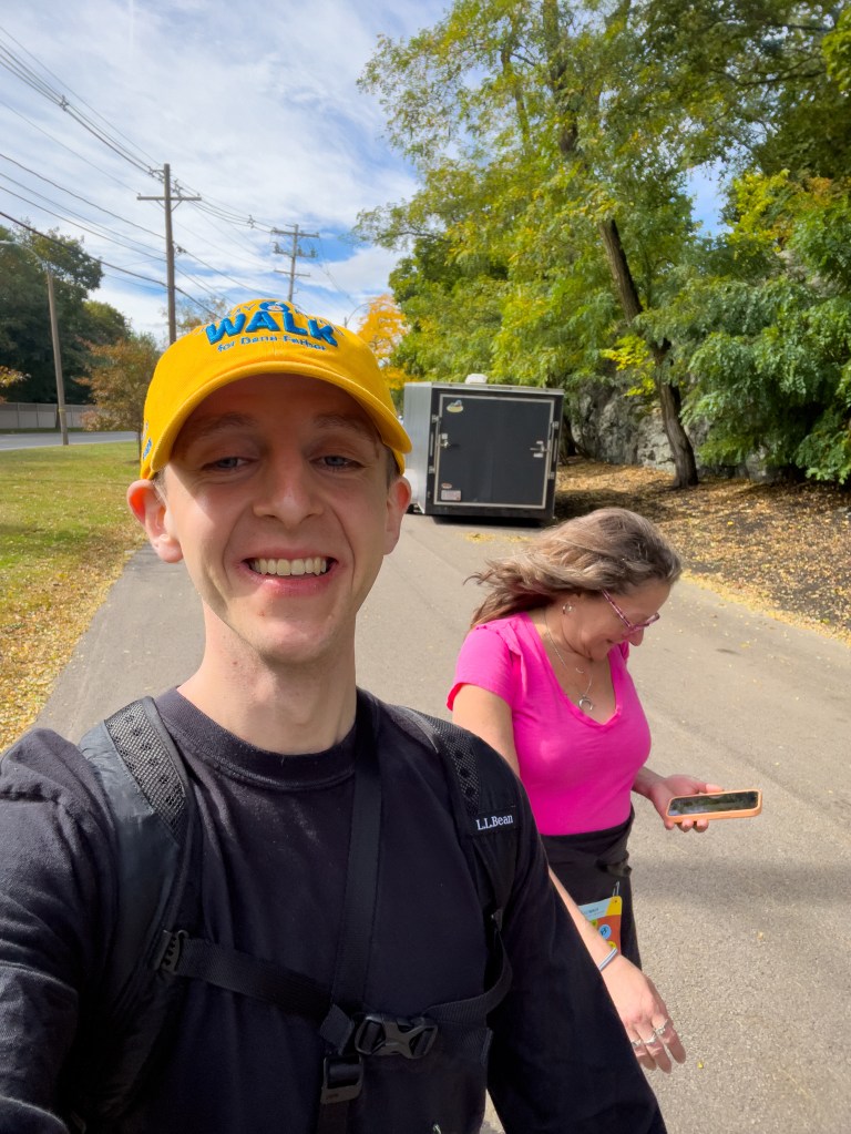 A young man smiling for a selfie while walking along a tree-lined path on a sunny day, wearing a yellow cap and black shirt. A woman in a pink top walks beside him, looking at her phone.