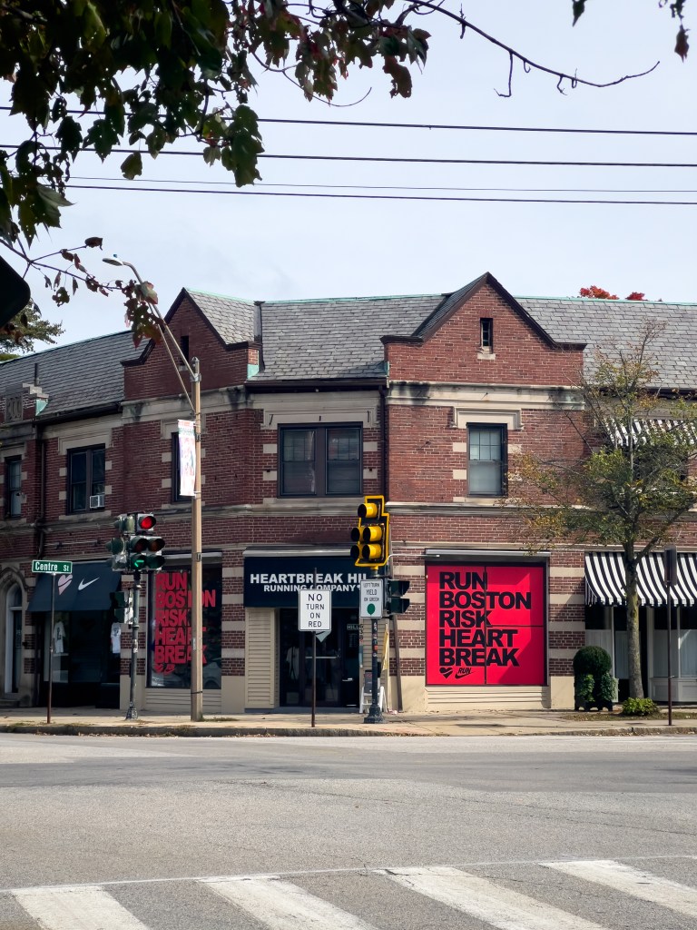 Street view of a brick building featuring a running store named 'Heartbreak Hill Running Company' with bold signage, located at an intersection with traffic lights and a nearby Nike store.